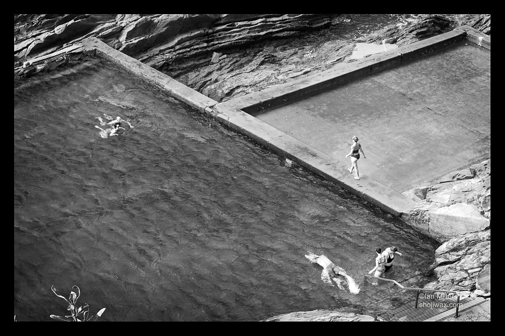 Black and white photo of ocean rocks swimming pool taken from above. There are people swimming and someone walking along one edge of the pools.