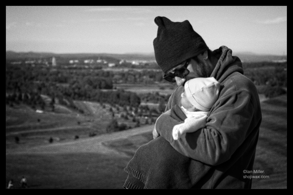 A man dressed in warm black clothes stands on a hill holding his new baby. In the background the city of Canberra can be seen.
