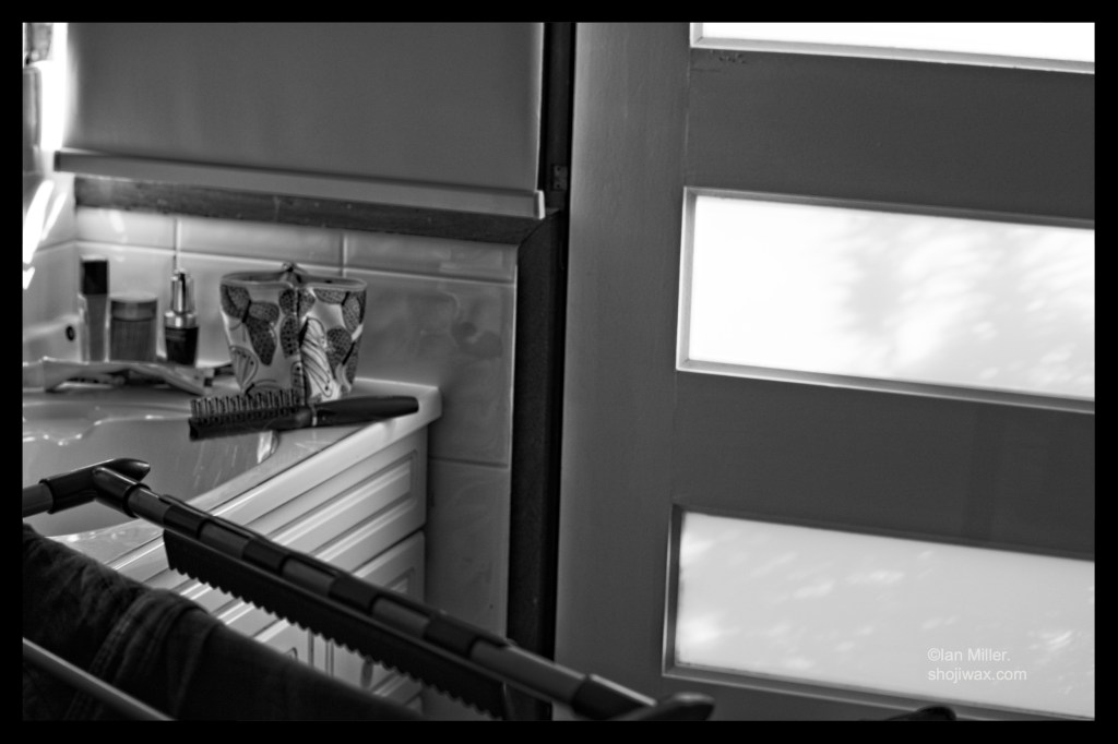 Black and white photo of a bathroom sink cluttered with personal hygiene items. There is a dappled light coming across from a door. In the foreground is a laundry rack.