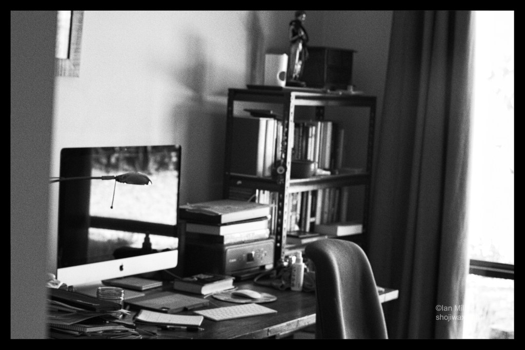 Black and White photo of a study.
There is a desk with a computer and various books and documents spread out on it . Next to the desk is a cluttered bookcase.
