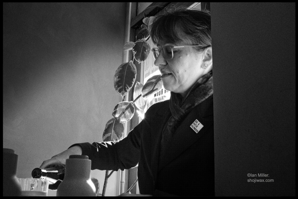 Black and white photo . Close up of lady in profile pouring water from a bottle. She is waring a dark top and a small white badge with the print: Be a nice human, on it.