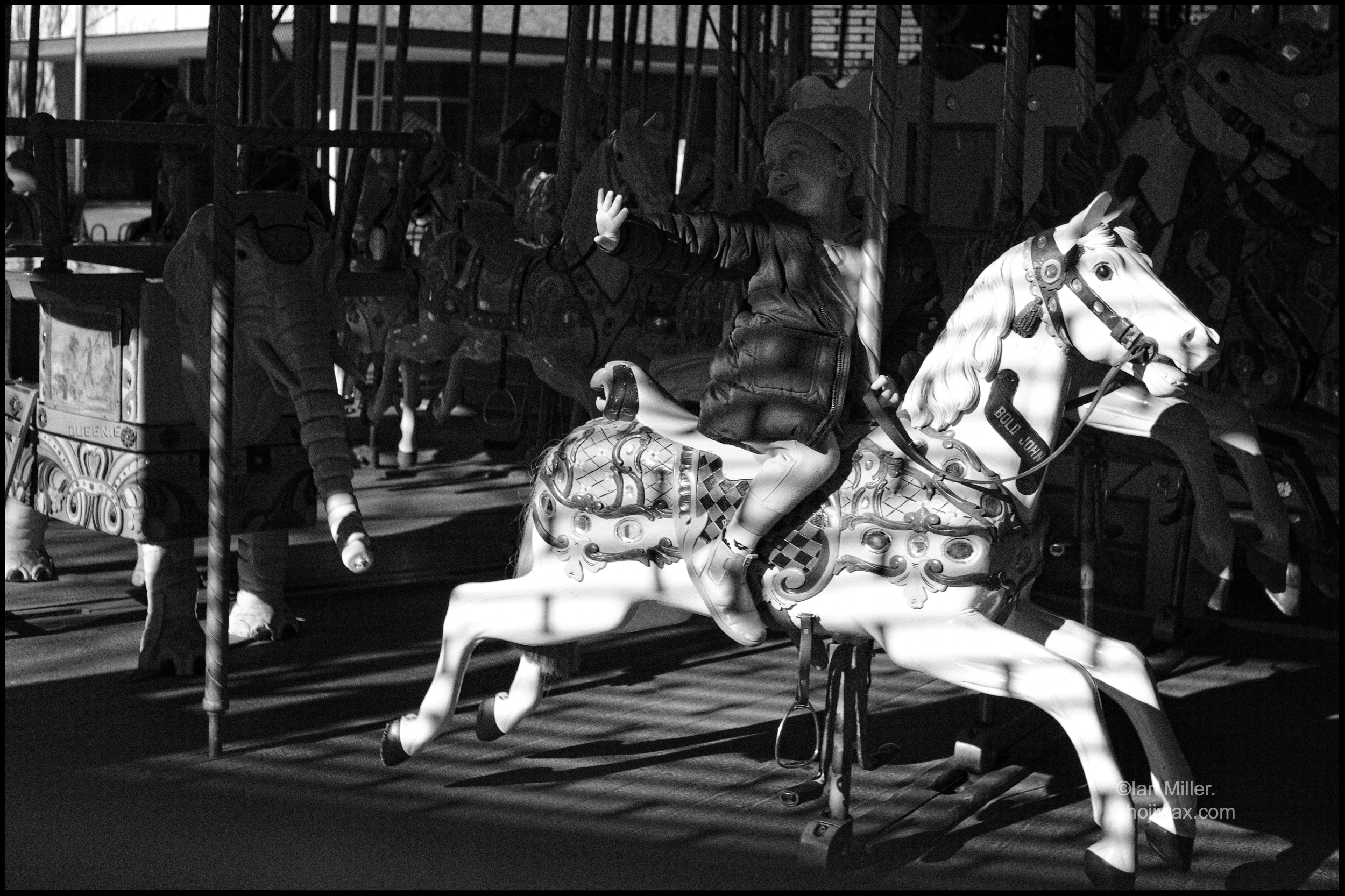 A merry-go-round in black and white. Most of it is in shadow except for a white horse in full light. On the horse a girl is also in shadow except her outstretched hand is in a shaft of light.