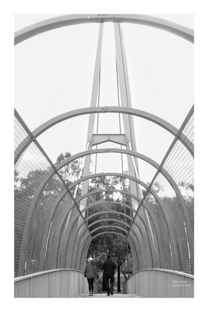 Black and white photo looking across an overpass. Two people are at the far end walking away from the camera. There are concentric metal arches above the overpass. The photo is overexposed.
