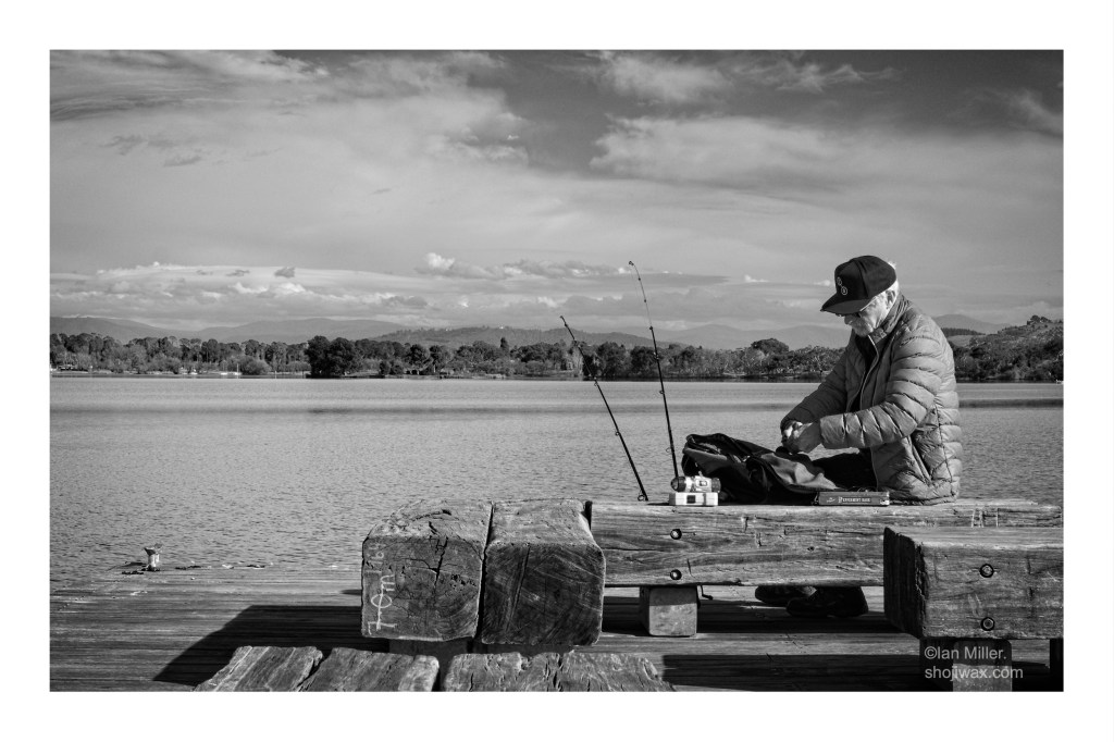 Black and white photo of an old man fishing at the end of a jetty. He is attending to his line whilst two fishing rods are seen beside him. Behind him is a large lake.