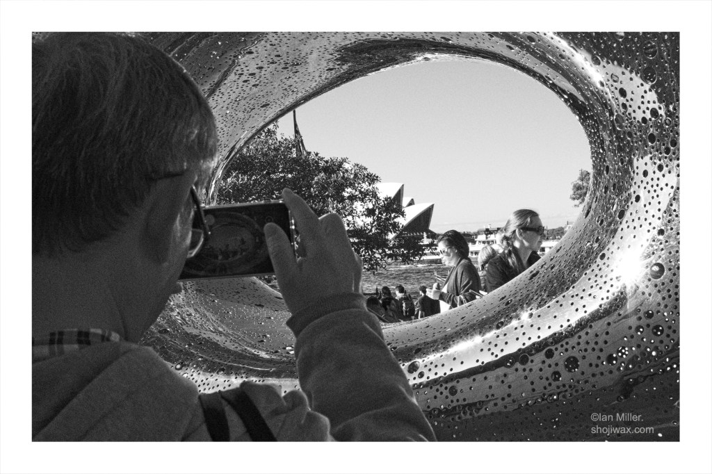 Black and white photo of man taking a phot through a elongated stainless steel doughnut shaped sculpture.