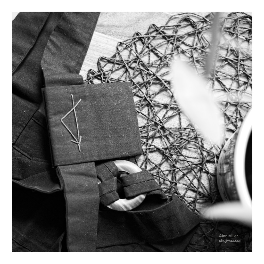 Black and white photo of a rakusu taken from above. It is sitting crumpled on a wooden table with a potted plants partially in view.