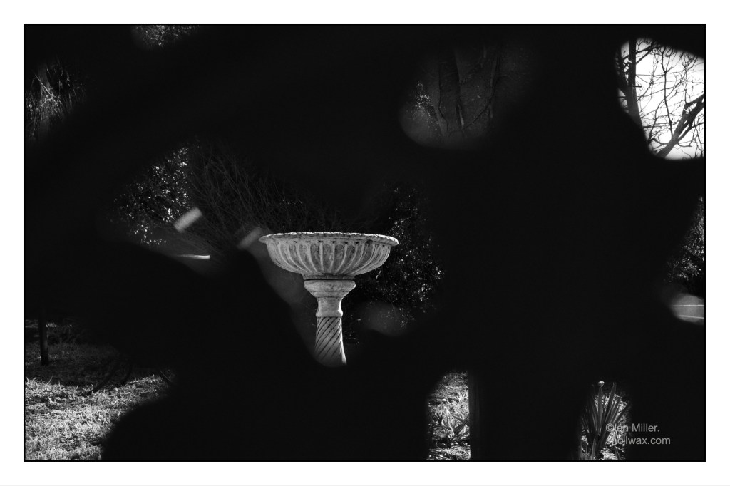 Black and white photo of a concrete birdbath seen fragmented behind deep shadows of an unknown object.