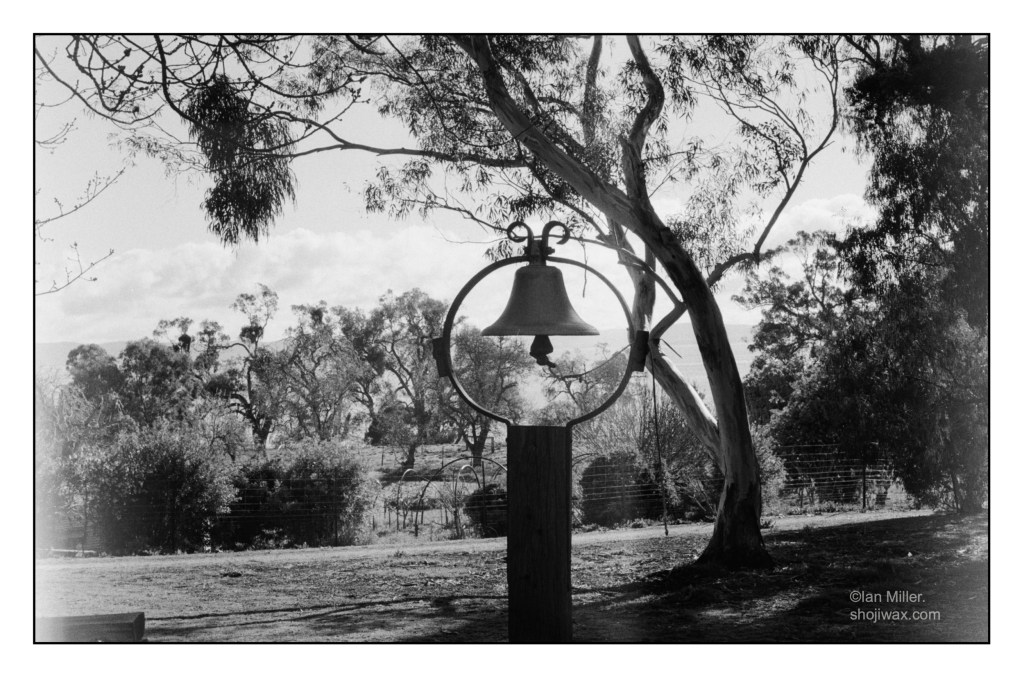 Black and white photo of a small brass bell atop a post and supported by an iron ring. In the background is an australian bush scene.