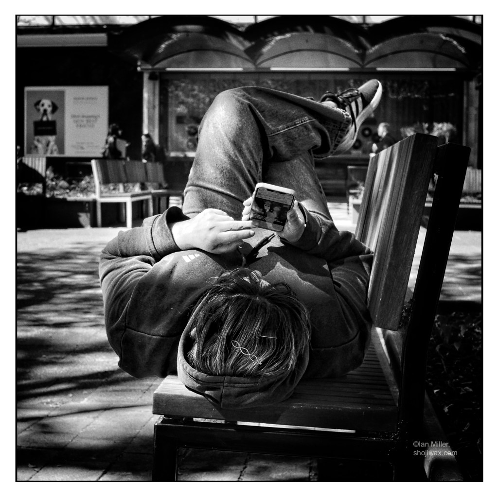 Black and white photo of girl laying on park bench checking her iPhone.