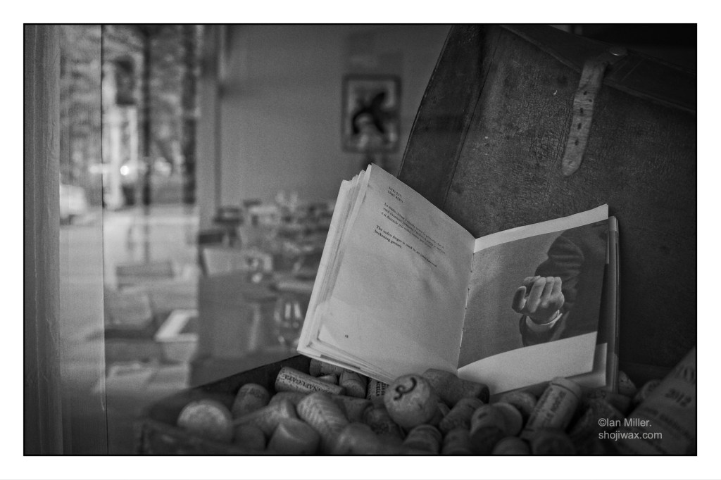 Black and white photo of shopfront display featuring an open book sitting on a bowl of corks. The photo in the book is of a hand making a beckoning gesture.