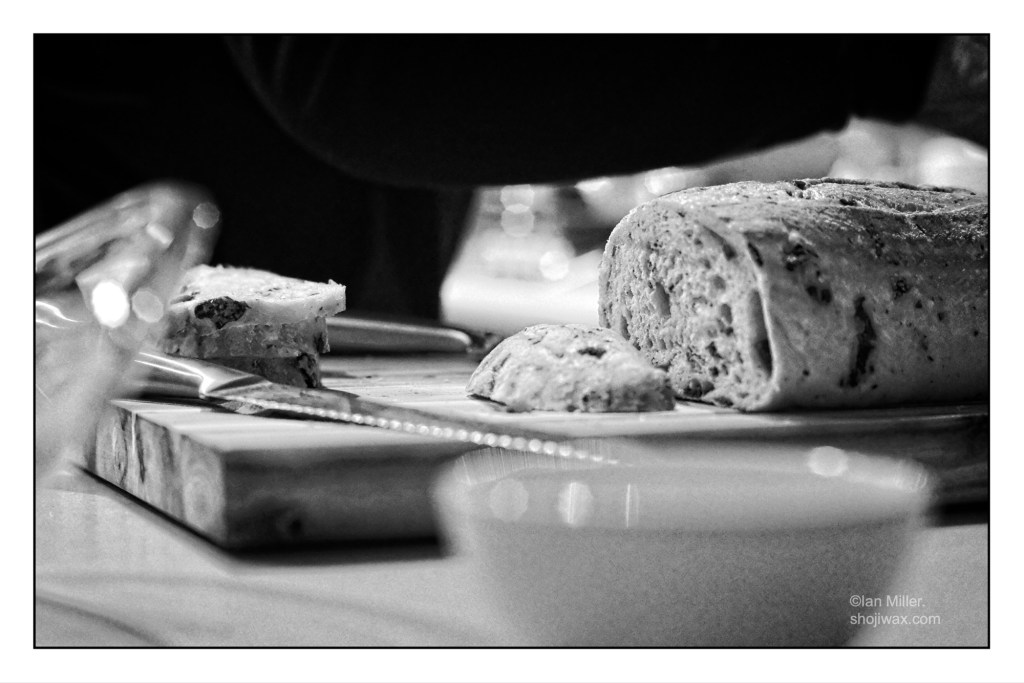 black-and-white photo of a loaf of bread on a wooden chopping board. There is a knife lying on the board and several slices of sliced bread. The background scene is of a kitchen but is out of focus.