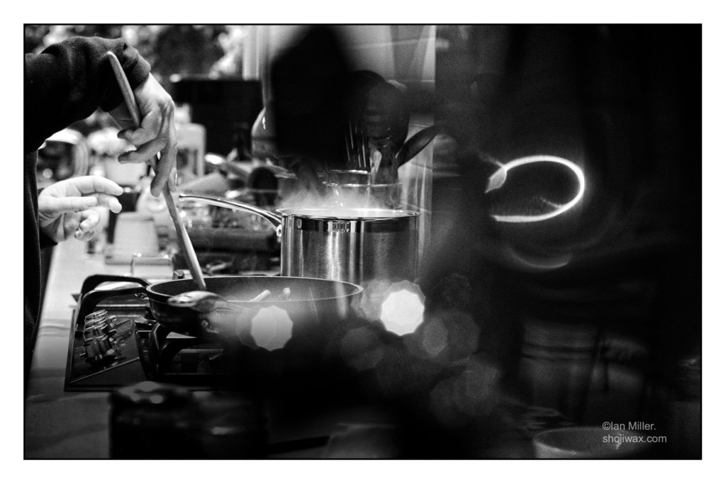 black-and-white photo of kitchen stove with metal pot and saucepan. There is lots of stainless steel. On the left of the photo a woman’s hand can be seen holding a wooden spoon..