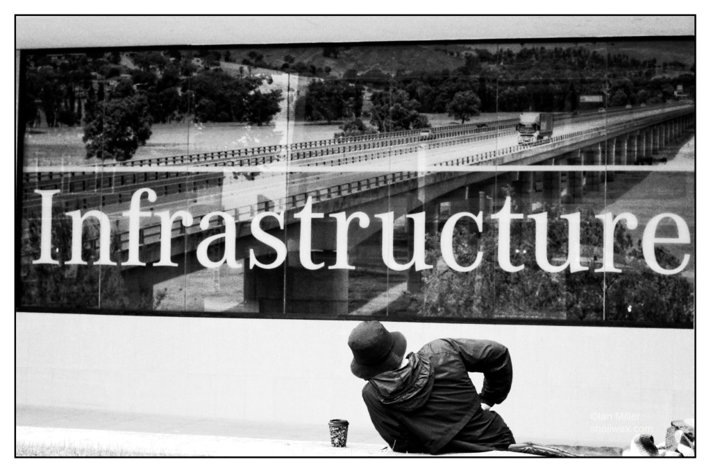Black and white photo of man half-laying underneath a large sign that says "Infrastructure"