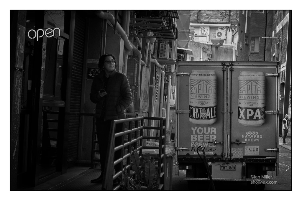 Black and white photo of alleyway. On the left side half hidden in shadows is a lady waiting at a small window for her coffee. There is a neon OPEN sign above her. Parked in the alleyway is a beer delivery truck.
