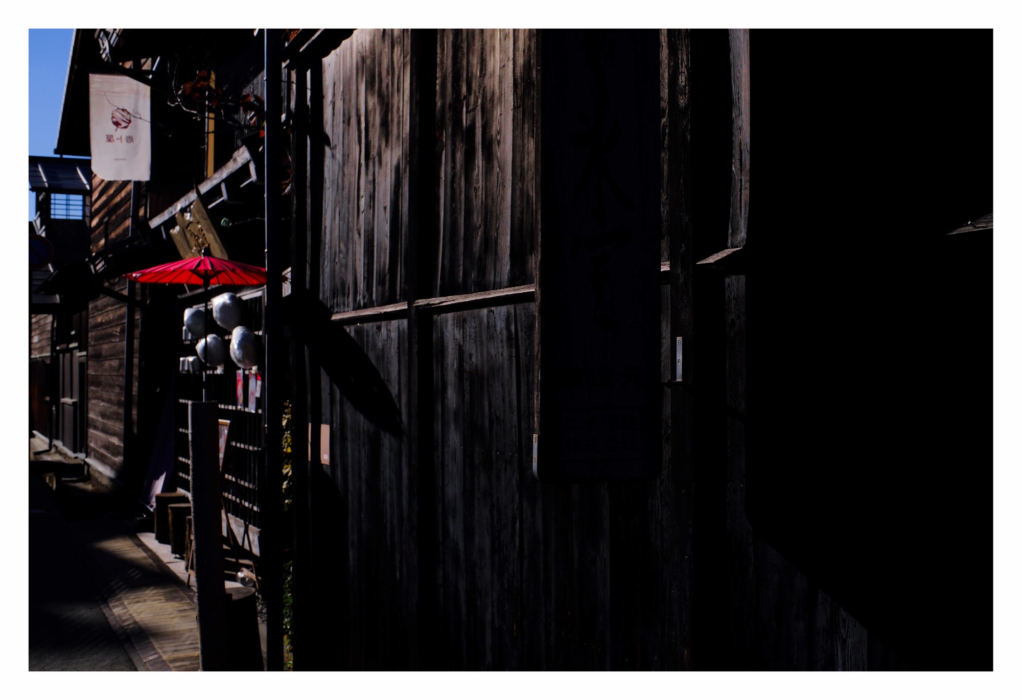 Japanese street scene. Predominantly dark wooden buildings with a single bright red inverted parasol on the left side of the image.