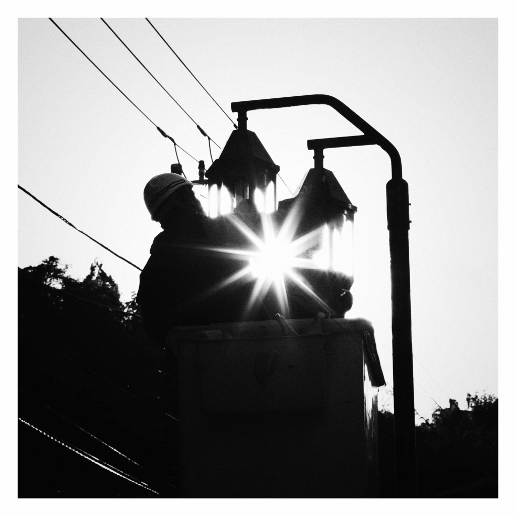 Black and white photo of a man silhouetted against the sun as he cleans a set of streetlights.