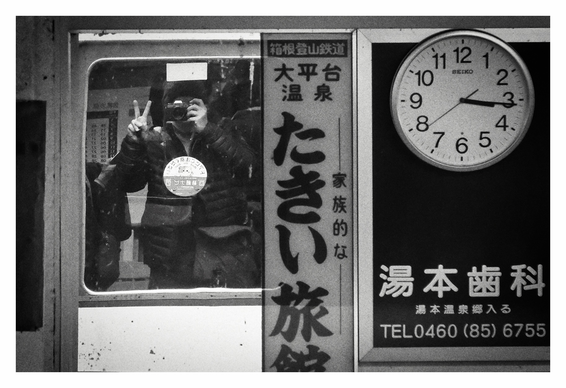 Black and white photo reflection of me through a train window holding camera up to my eye. Beside me is a large station clock. I am making a peace sign.