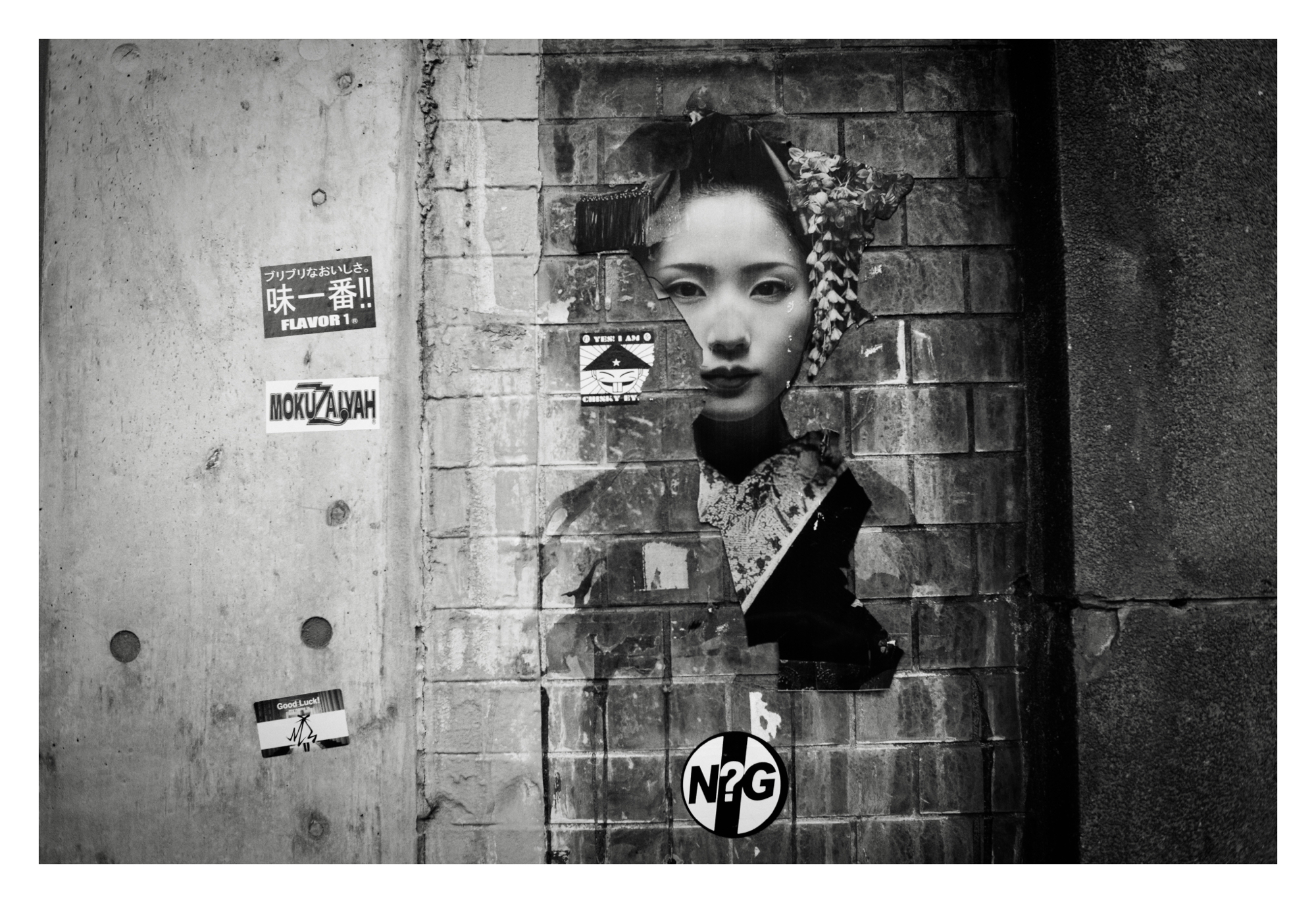Black and white photo of a torn poster depicting the face of a Japanese woman. There are various stickers on the wall surrounding the poster.
