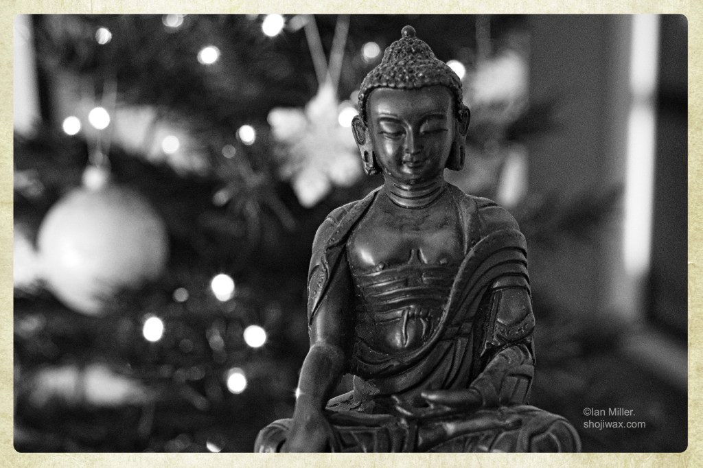 Black and white photo of a wooden Buddha statue in front of a lit christmas tree.