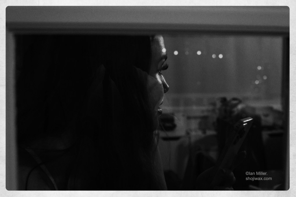 Black and white photo. Very dark image taken through a window of a womans face in profile and lit from one side.