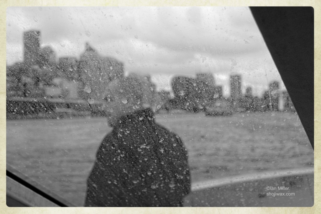 Black and white photo shot through a dirty window. Out of focus man looking over the bow of a ferry at the distant city skyline.