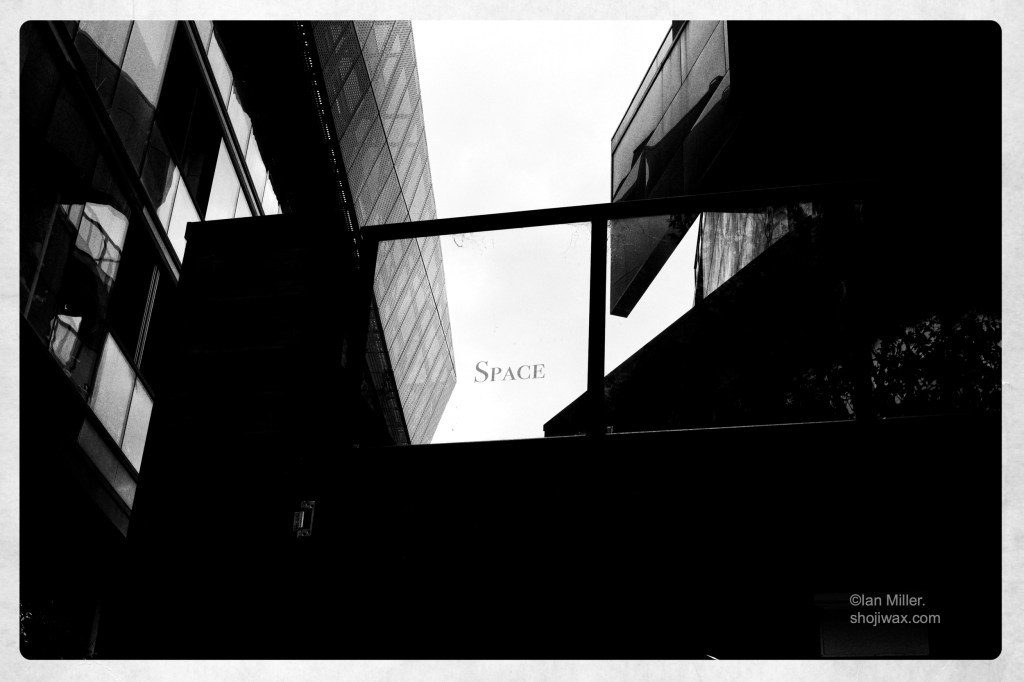 High contrast monochrome photo of two adjacent buildings with a window in between. Written on the window is the word: space.