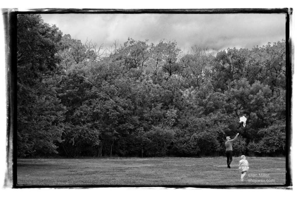 Monochrome photo of man and small girl running with a kite towards a long stand of trees.