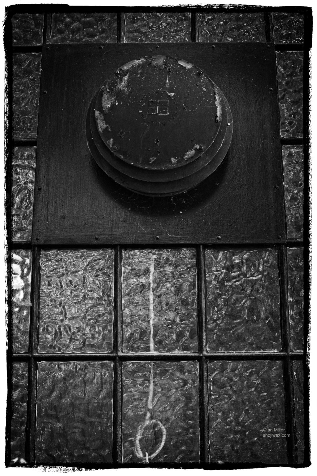 Monochrome photo of old window with air fan in the upper portion. The glass is old and has a scalloped finish. There is an electrical cable visible hanging down behind the glass.