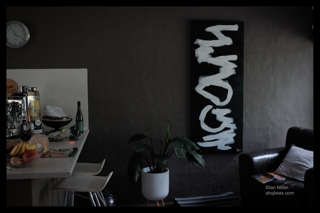 rectangular painiting of simple bold white squiggles on a black background. Around the painting are a large leather chair, a pot plant and an espresso machine on a bench.