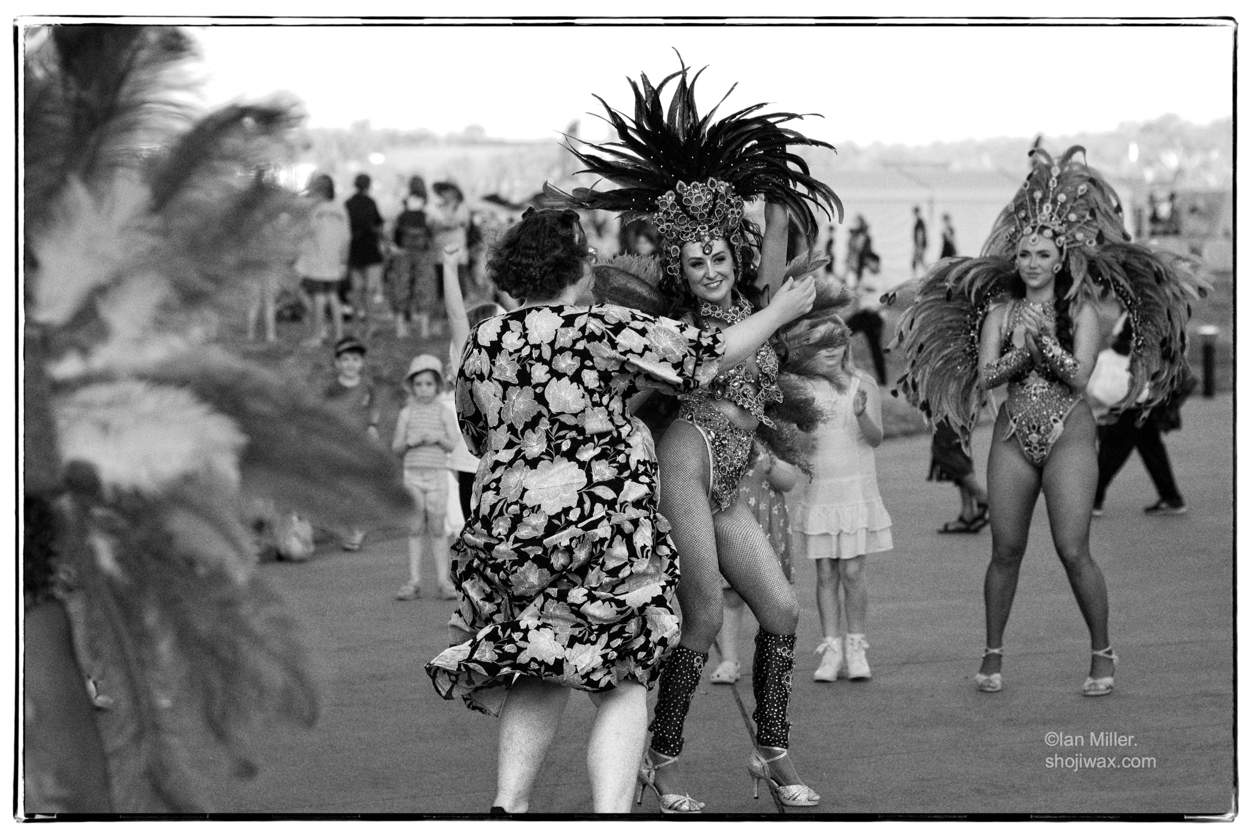 Monochrome photo of young lady dressed in feathered costume dancing with older lady in a floral plain dress.