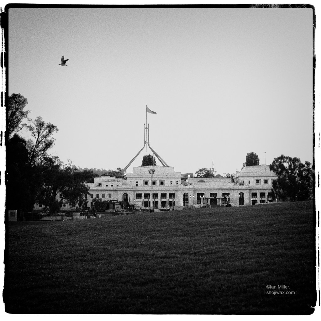 Black and white grainy photo of old white building. Behind and in the distance is a modern large flagpole.