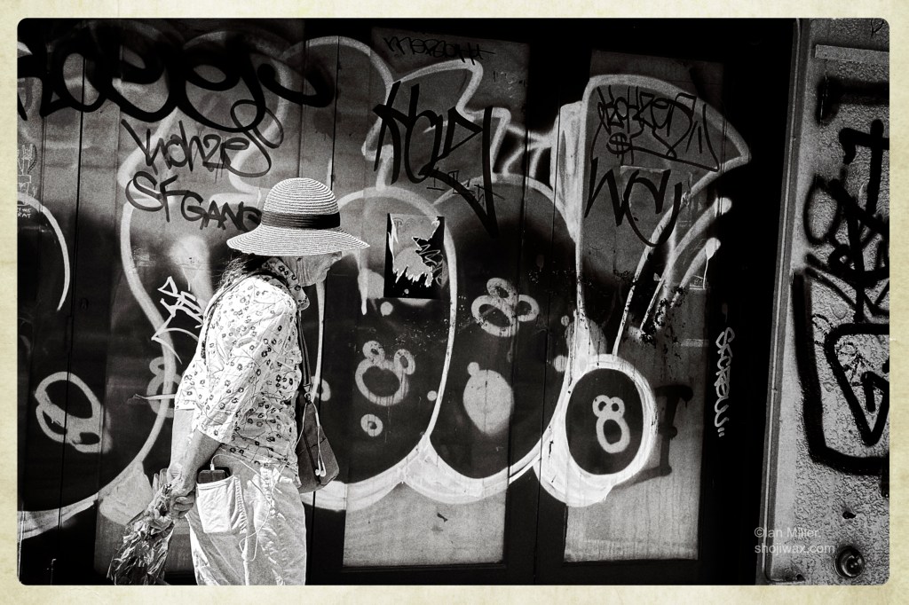 Black and white photo of older woman wearing a sun hat walking along holding a bunch of flowers. The background is a wall of high contrast graffitti.