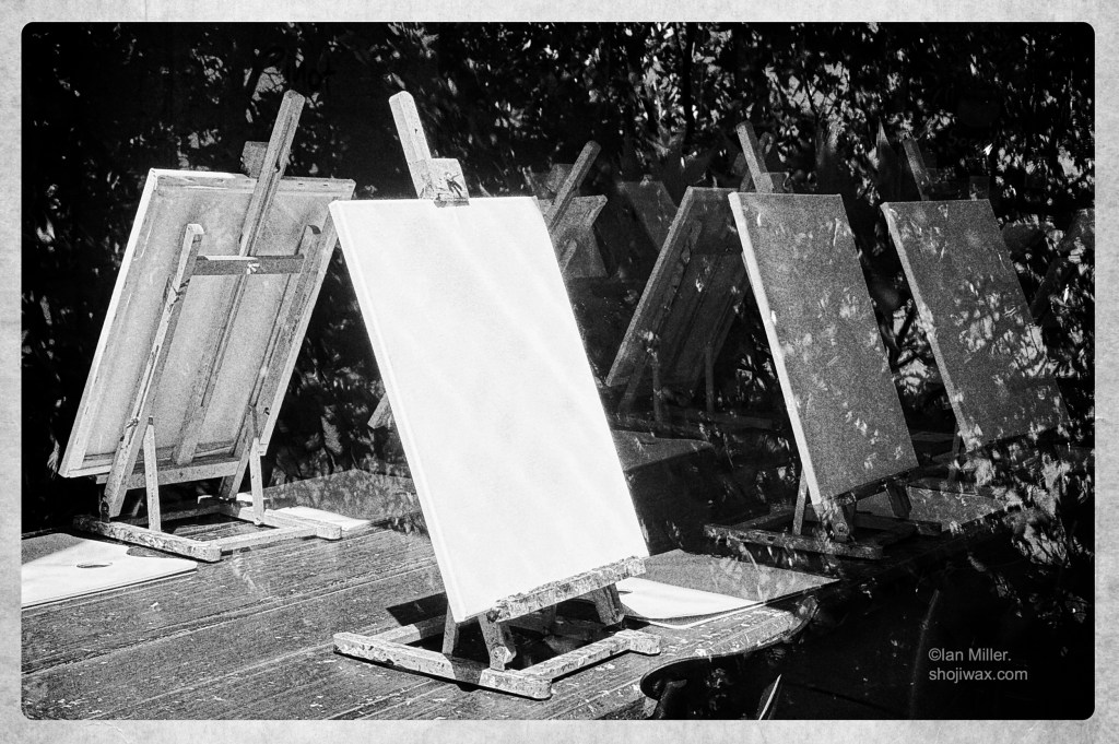 Black and white photo of row of easels on a wooden table. Each one holds a blank canvas. The photo is taken through a window and there is a reflection of tree branches.