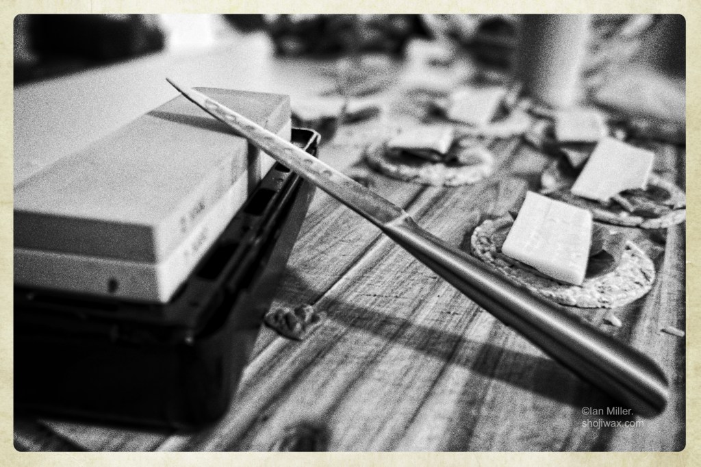Black and white photo of a whetstone sitting on a wooden cutting board. There is a knife leaning against it and in the background are cheese and crackers.