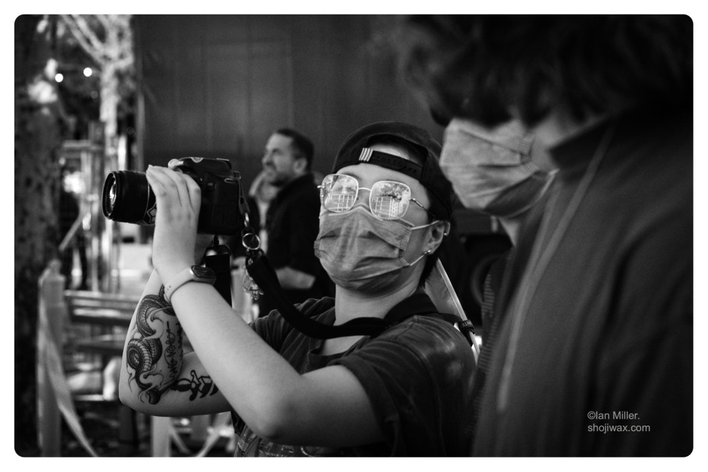Monochrome photo of female holding a camera. She is wearing a cap and surgical mask.