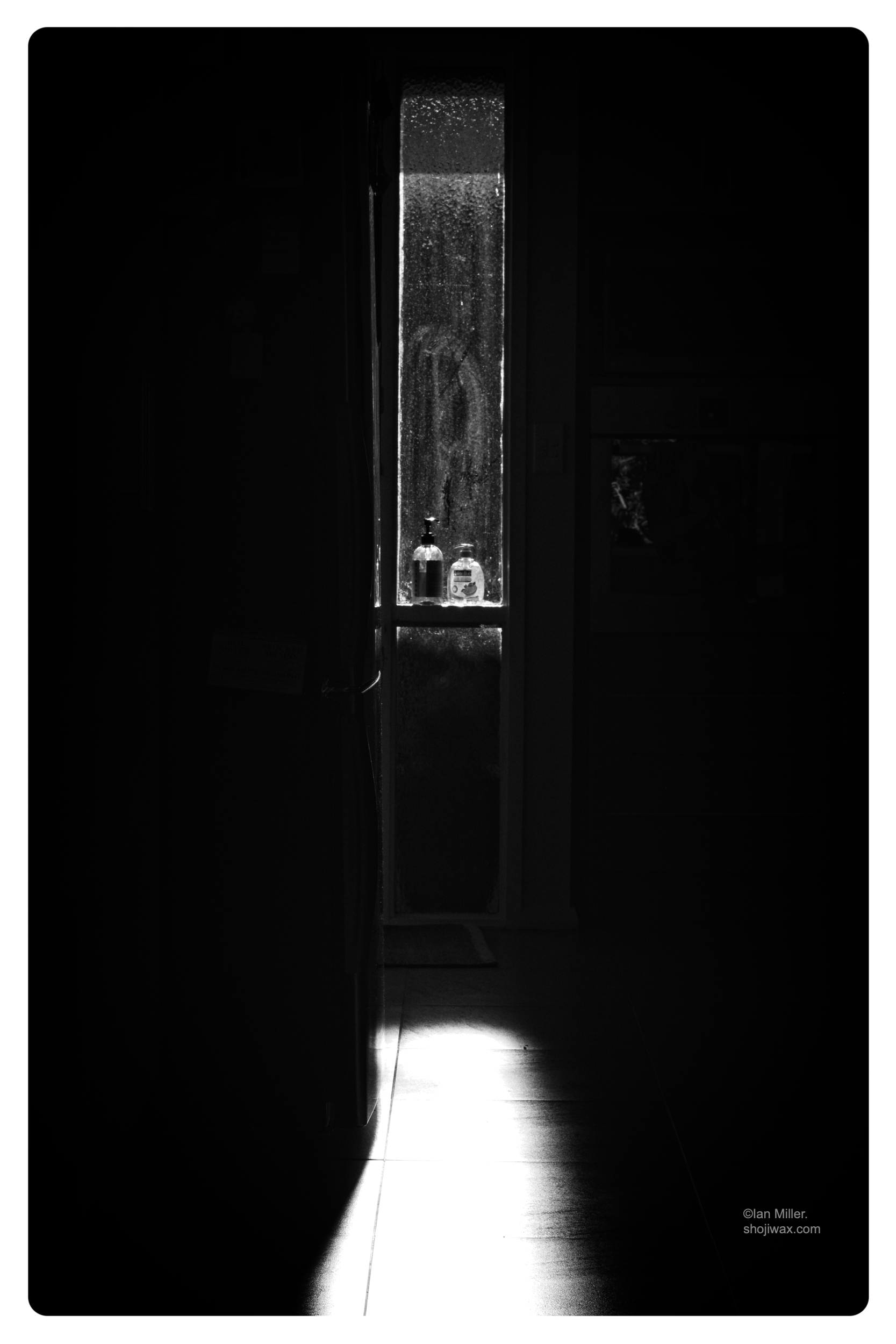Monochrome high-contrast photo of a narrow vertical window with light streaming through casting light on the floor. In the centre of the window are two small soap dispensers.