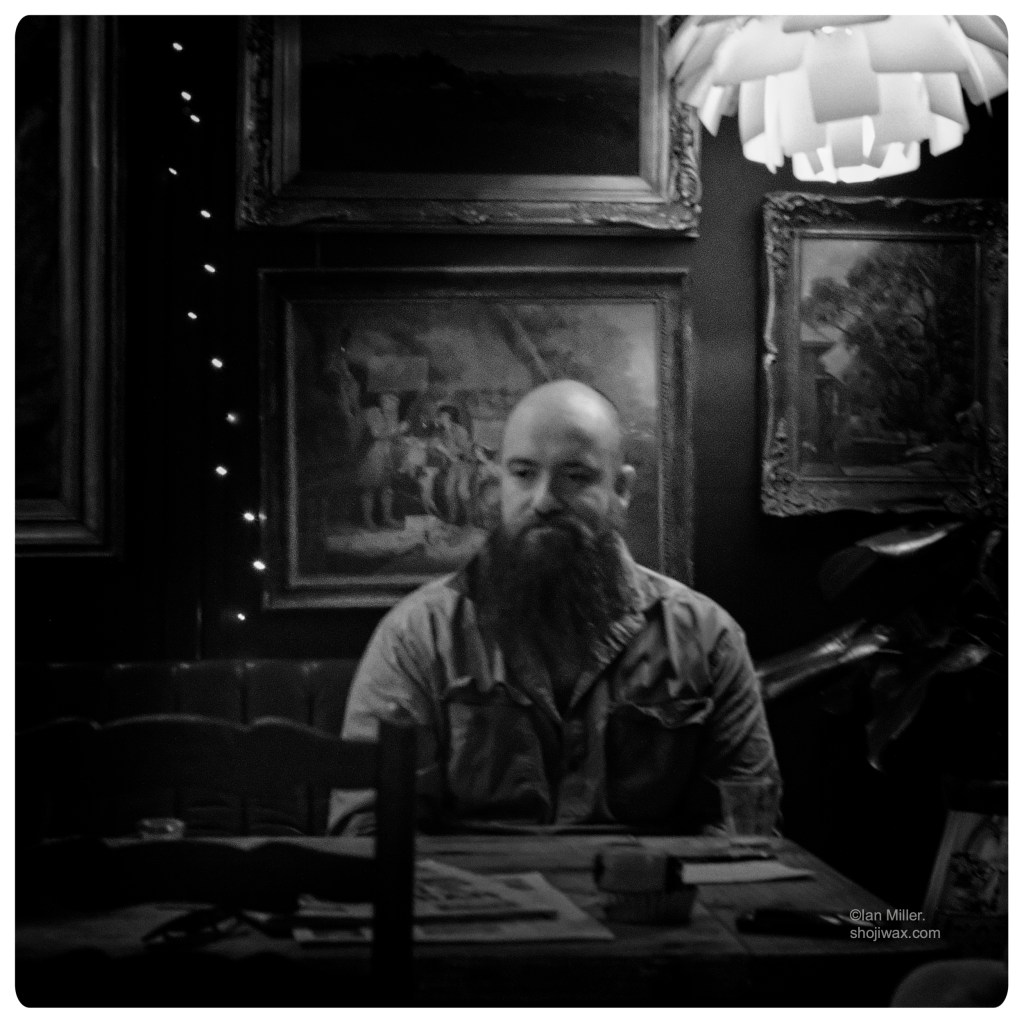 Monochrome photo of a bearded, bald man sitting at a table surrounded by old paintings on the wall behind him.