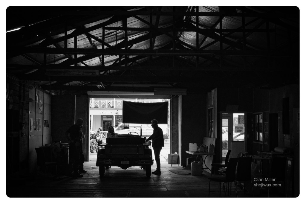 Monochrome photo of an old vintage car seen in silhouette in front of the entrance to a large old garage/workshop.