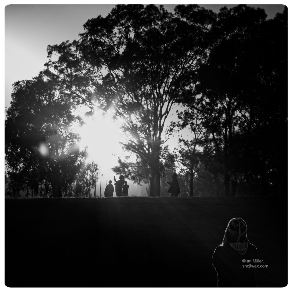 Monochrome photo of two people silhouetted against a sunrise through trees. Another person can be seen in the foreground.