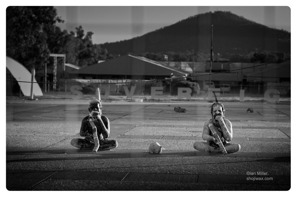 Two small statues of first Australians sitting playing the didjeridoo. In the background is a large sign saying 'sovereignty'