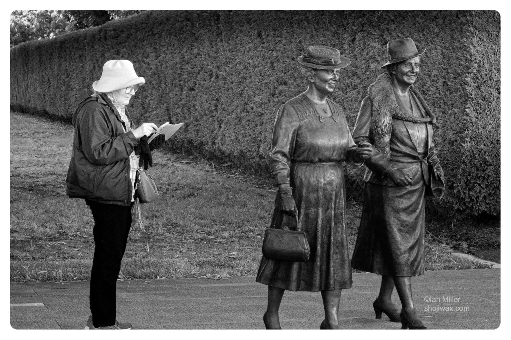 Monochrome photo of elderly woman wearing a hat checking her phone next to two statues of elderly women wearing hats.