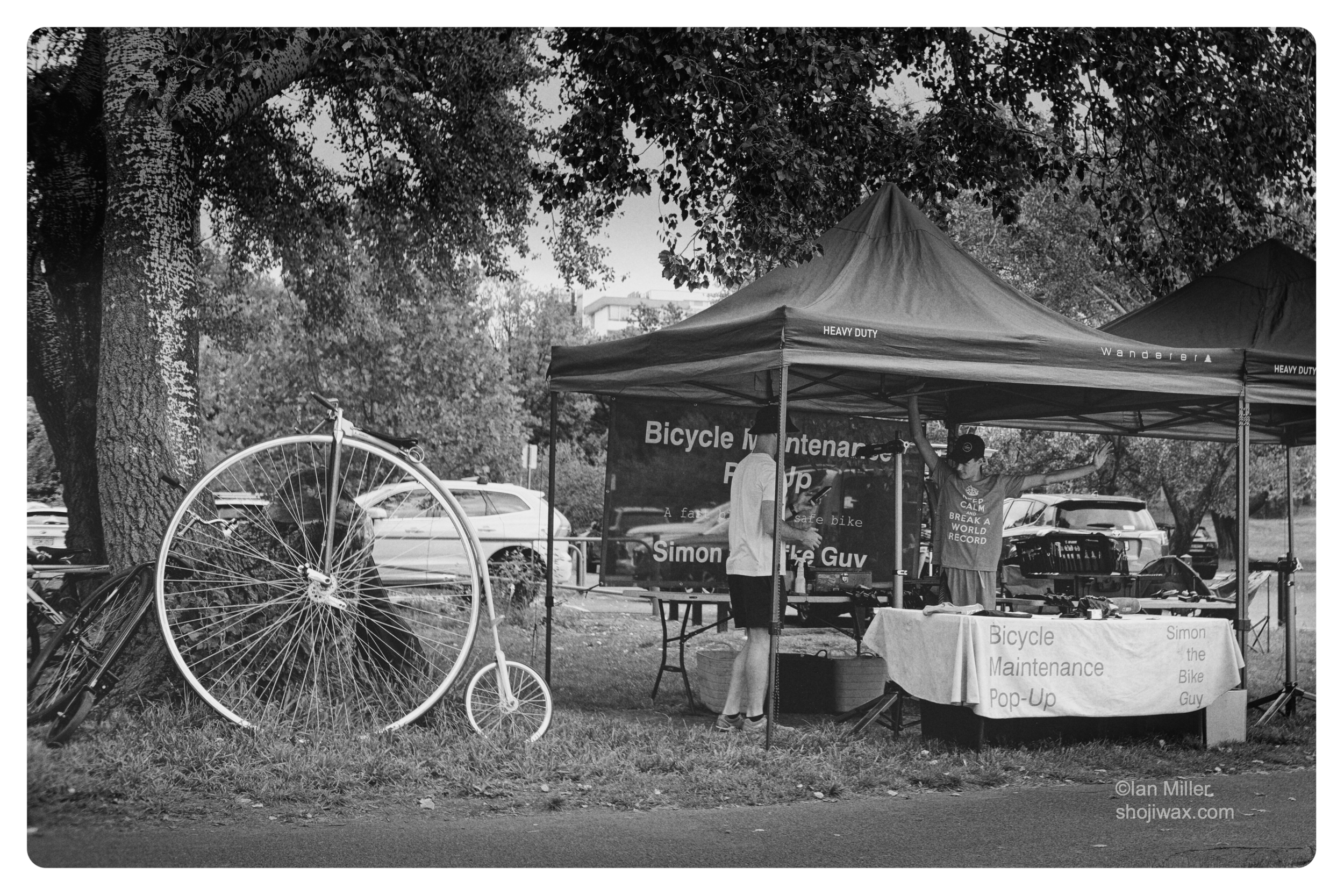 Monochrome photo of pop up tent housing a bicycle repair station. Beside the tent two Penny Farthing bikes rest against a large tree.