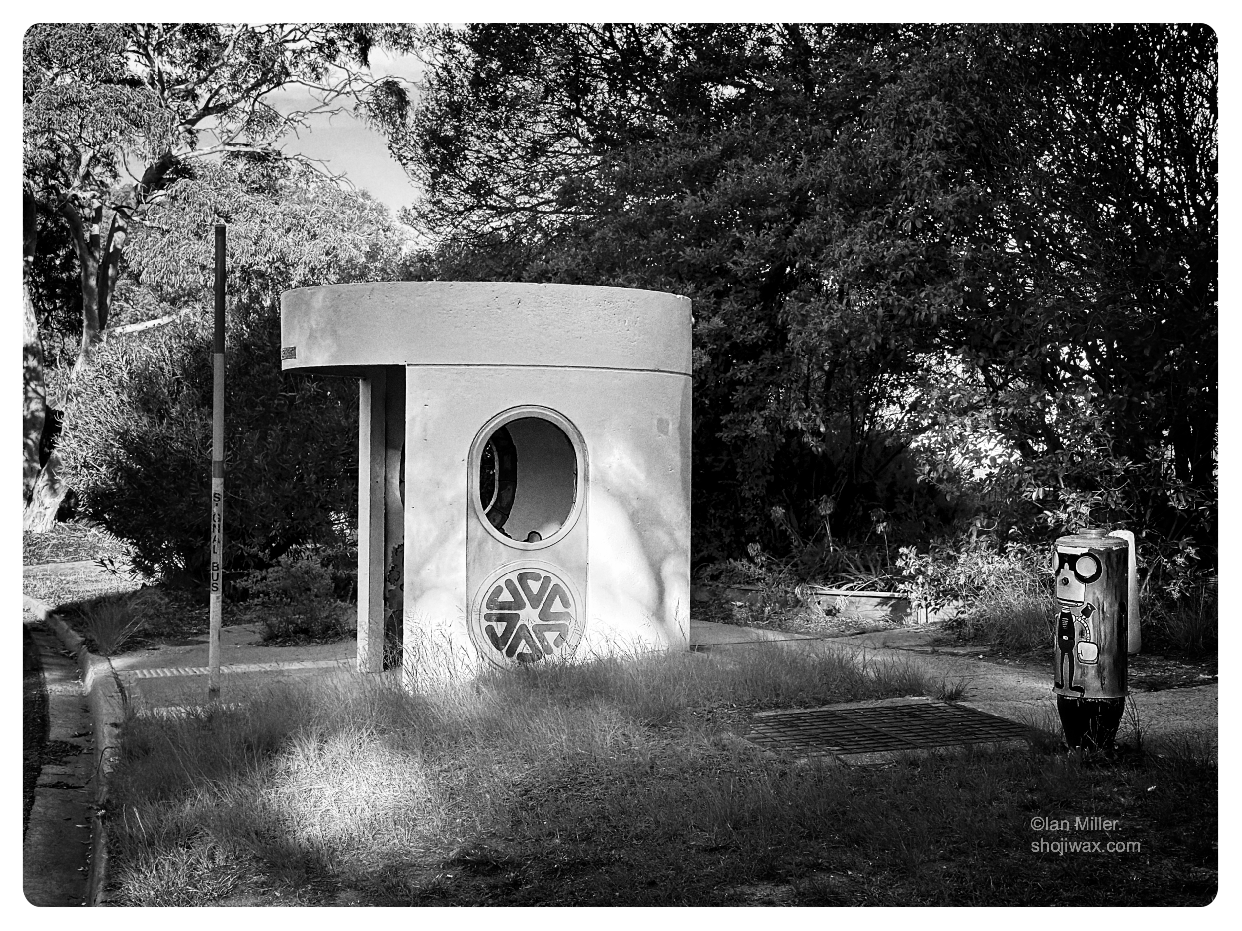 Monochrome photo of large cylindrical concrete bus shelter on a suburban street.