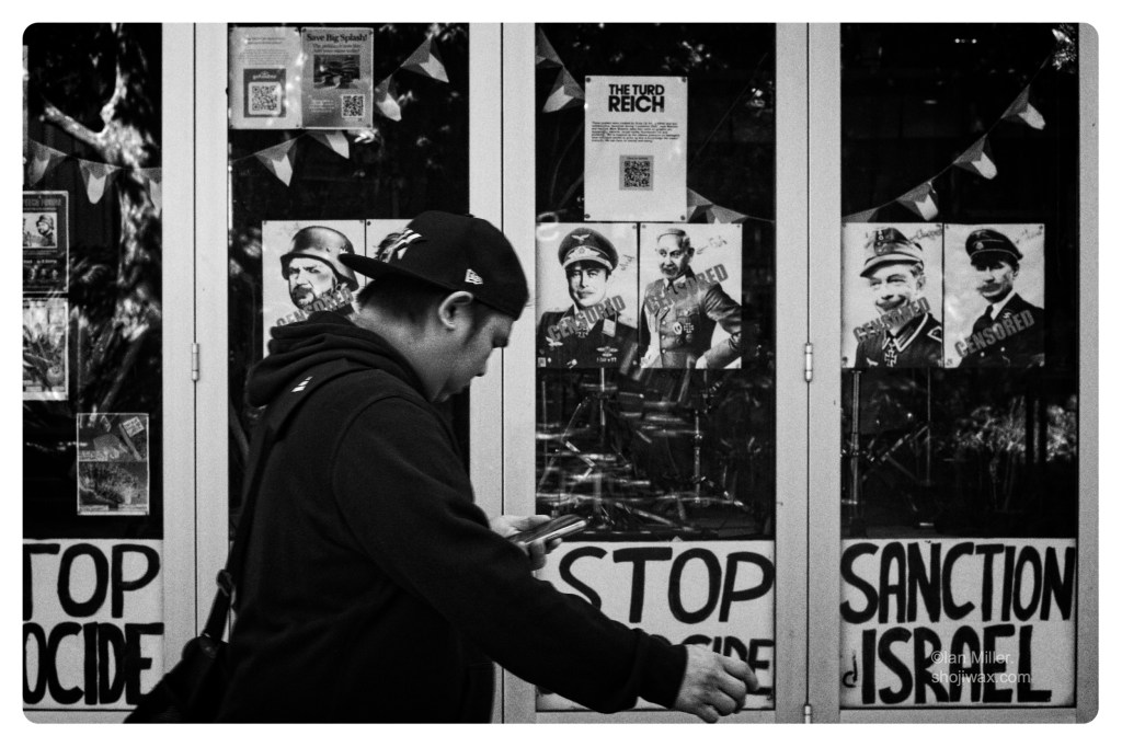 Monochrome photo of man walking by looking at his smartphone. In the background there is a window with protest posters.
