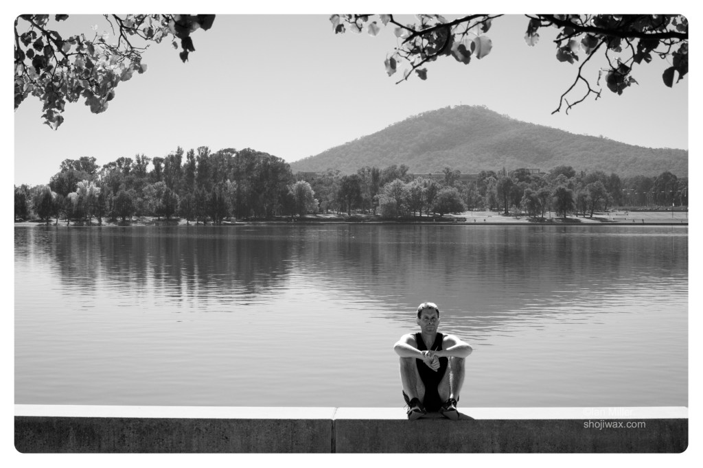 Monochrome photo of man sitting on a concrete wall besides a lake. In the background is the lake and a mountain and trees on the other side