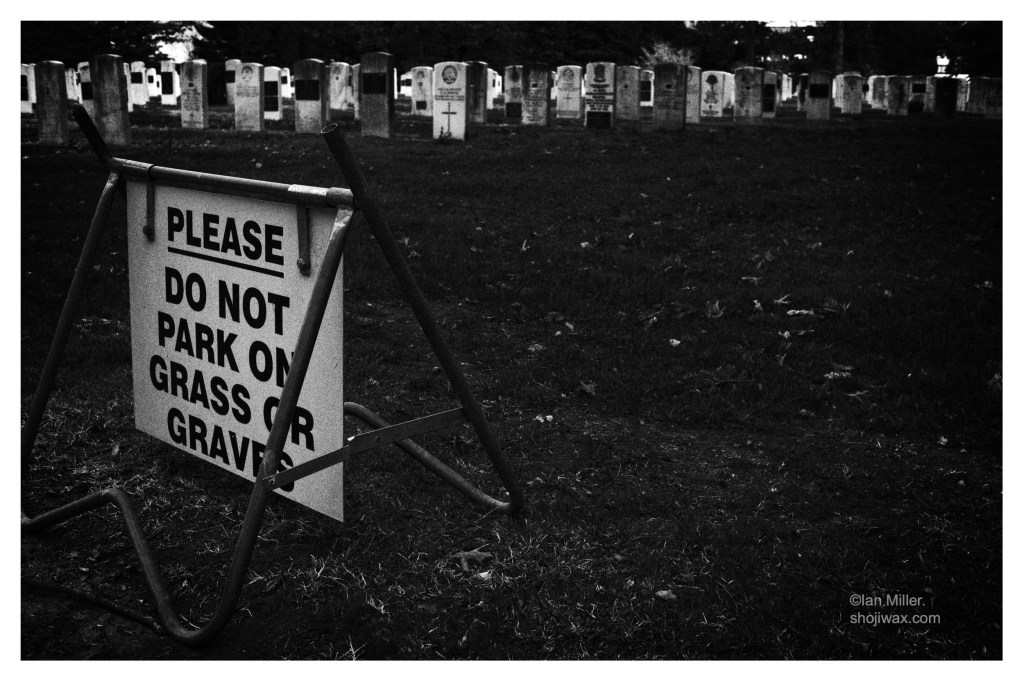 Monochrome photo of cemetery gravestones. A sign in the foreground says do not park on grass or graves.