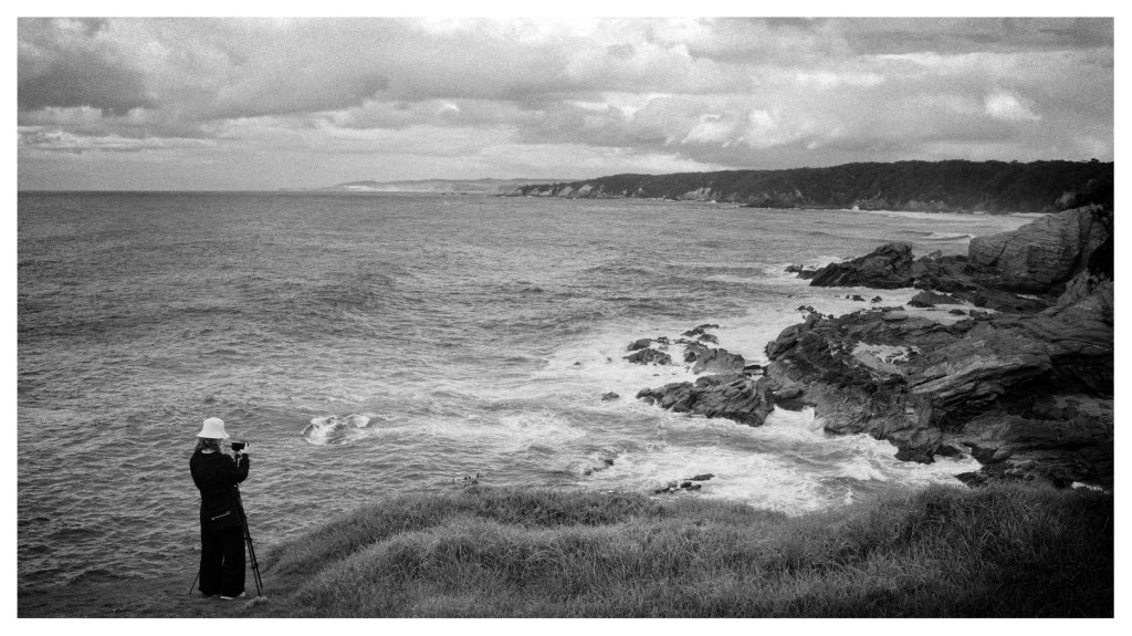 Monochrome photo of lady on a hill overlooking cliffs and a wild sea. she has a camera on a tripod.