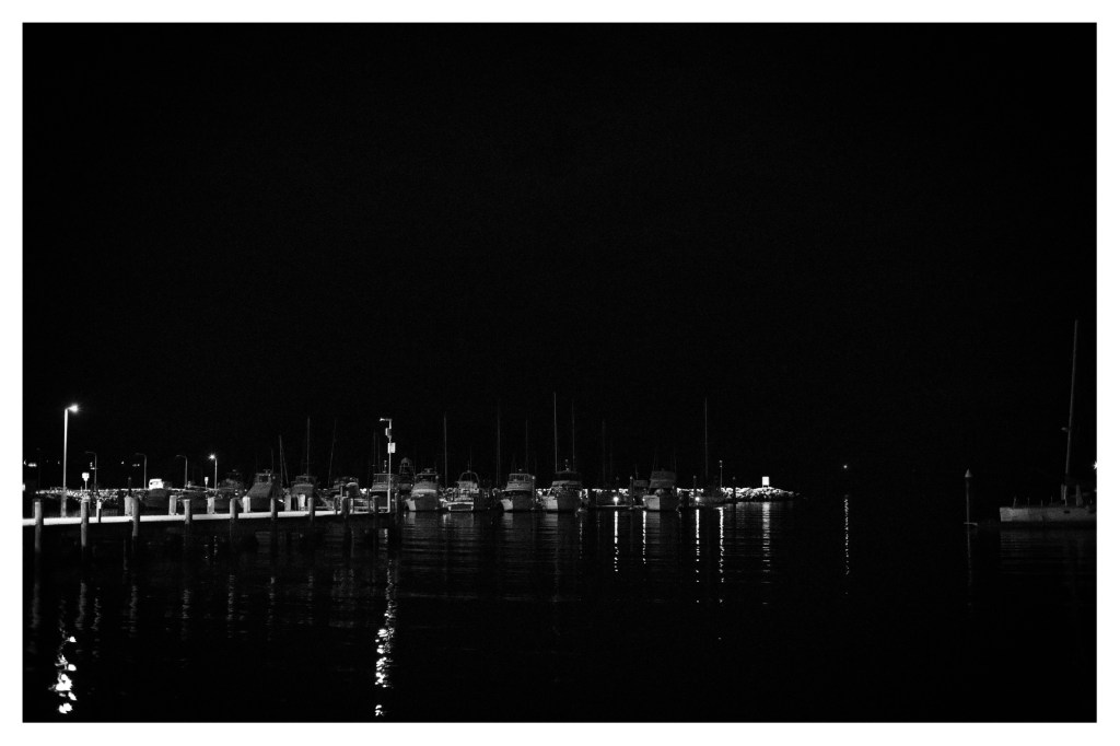 Monochrome nighttime photo of distant jetty and a long line of boats just visible along its length.