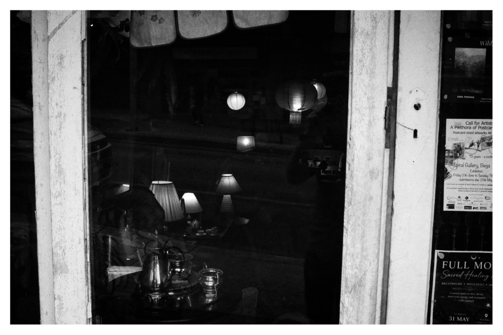 Monochrome photo take through a store window. There are vintage hanging lamps and a silver tea set visible in the darkness.