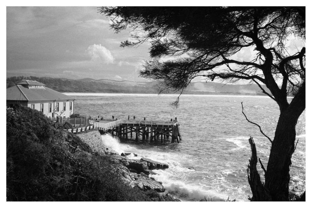 Monochrome photo of a distant wharf and building. People can be seen fishing on the wharf.